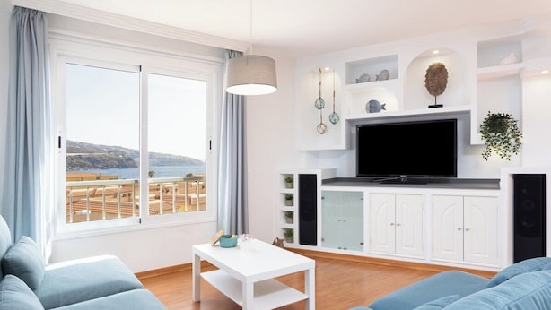 Ett modernt vardagsrum med ett stort fönster, en platt-TV, vita skåp och en blå soffa.