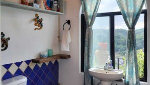 Une salle de bain avec des toilettes, un lavabo et des étagères au-dessus des toilettes. Il y a une fenêtre avec des rideaux et une vue sur l’extérieur.