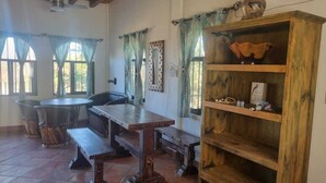A rustic dining area with a wooden table and benches, a wooden bookshelf with decorative items, and a window with a view of greenery. - Casa Pajaros - Balcony (La Cruz de Huanacaxtle)