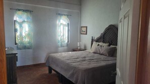 A bedroom with a bed, two windows with curtains, a bedside table with a lamp, and a door. - Casa Pajaros - Balcony (La Cruz de Huanacaxtle)