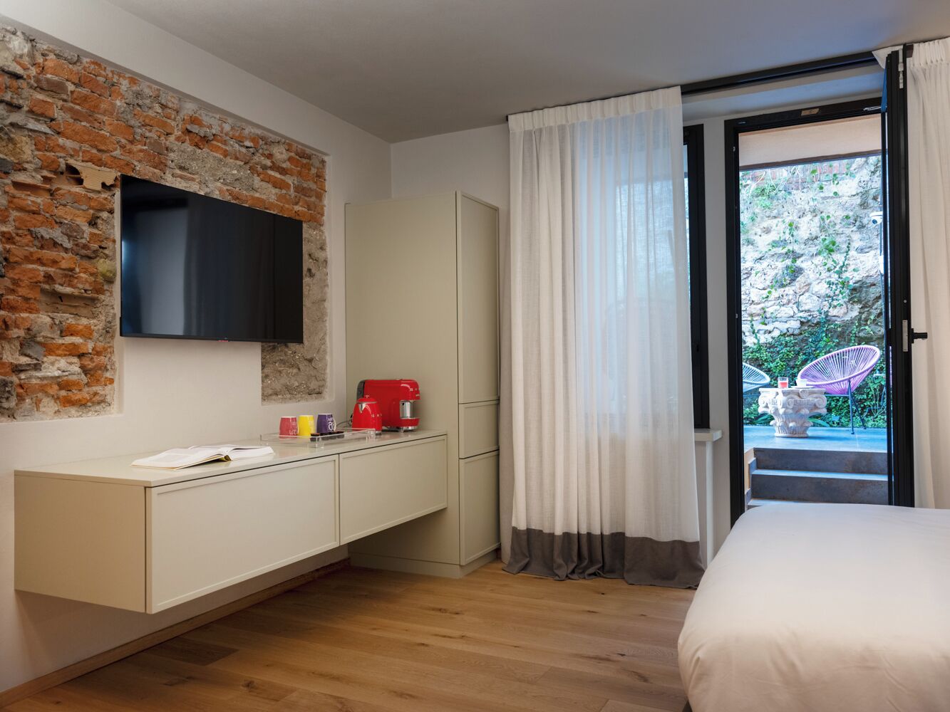 A modern room with a brick accent wall, a flat-screen TV, a white wardrobe, a red coffee maker, and a bed with a view of an outdoor area.