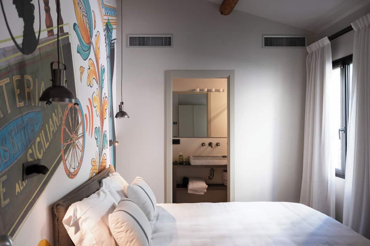 A bedroom with a bed, a nightstand, a mirror, a window with curtains, and a bathroom with a sink and mirror.