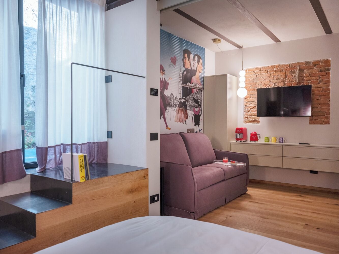 A modern bedroom with a sofa, a TV mounted on a brick wall, and a window with curtains.