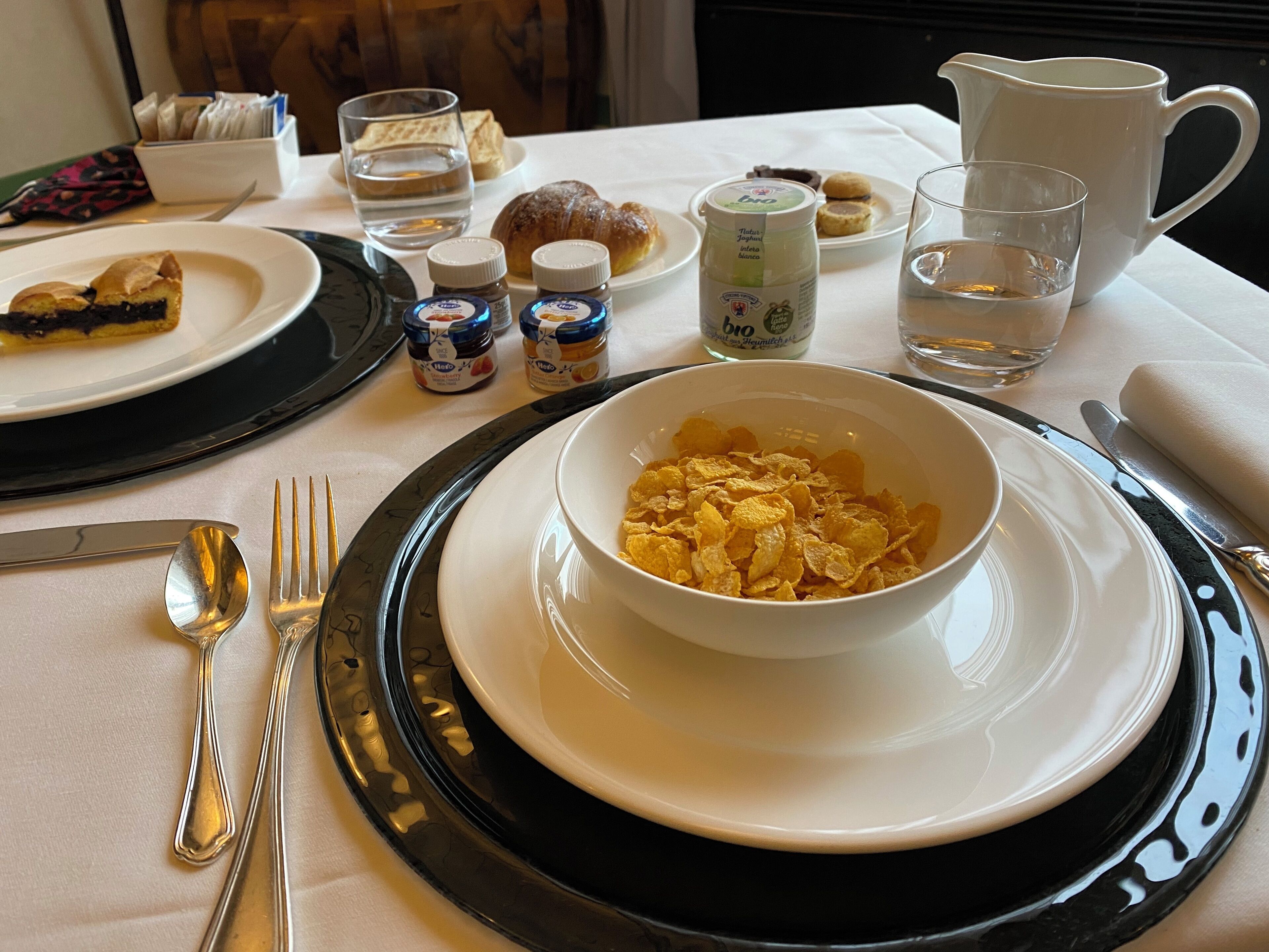 Una escena de desayuno con un tazón de cereal, un croissant y una jarra de leche sobre una mesa.