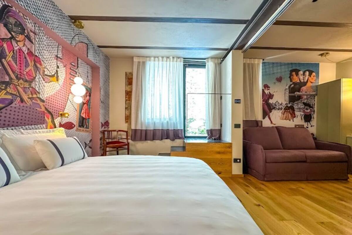 A hotel room with a large bed, a brown sofa, a mural on the wall, and a window with curtains.