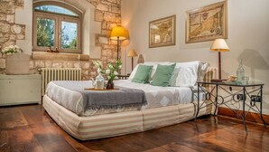 A bedroom with a stone wall, a wooden ceiling, a bed with striped bedding, a radiator, a window with a view, and two wall-mounted lamps. - Villa Ramunna 6, Emma Villas (Rosolini)
