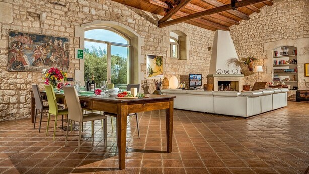 A spacious living area with stone walls, a wooden ceiling, a dining table with chairs, a fireplace, and a television. - Villa Ramunna 6, Emma Villas (Rosolini)