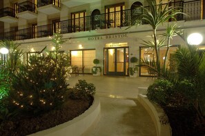 Hotel Bristol's entrance with a well-lit facade, a covered outdoor area, and a landscaped garden with illuminated trees. - Hotel Bristol (Alassio)
