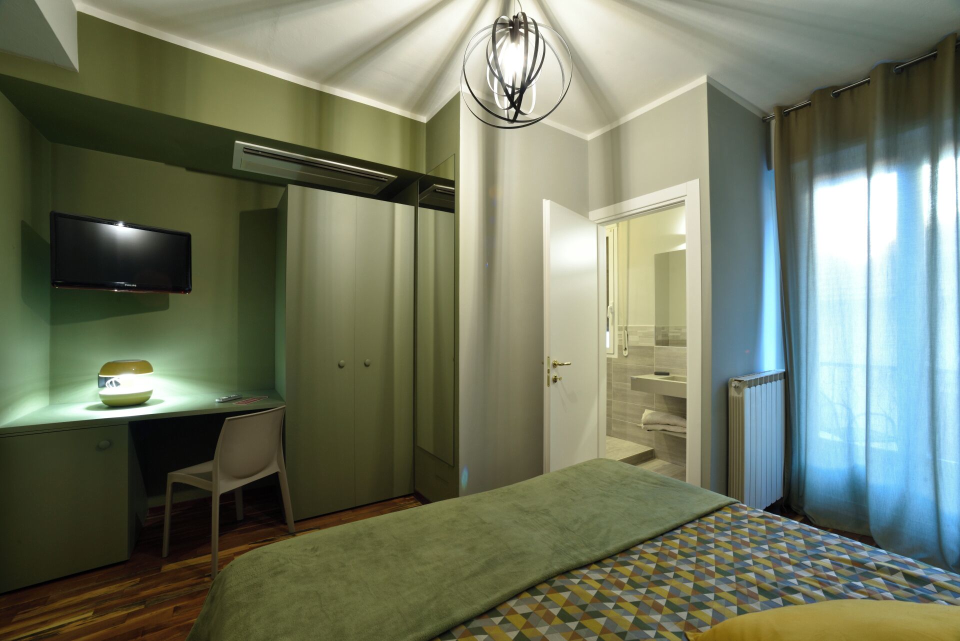 A hotel room with a bed, a desk, a chair, a television, and a bathroom visible through an open door.