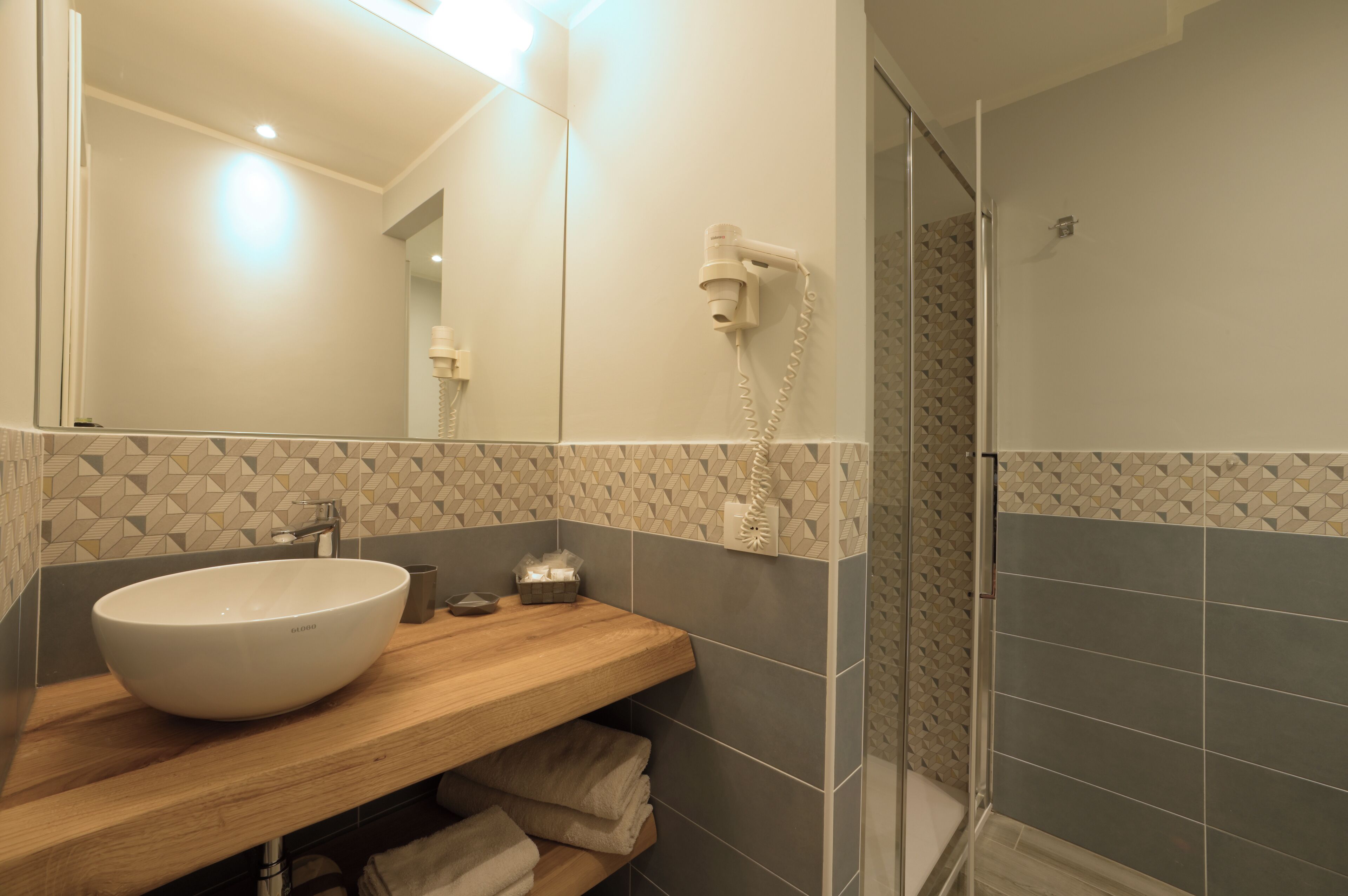Ein Badezimmer mit einem großen, weißen Waschbecken, einer Holzablage, einem Handtuchhalter und einer Duschkabine mit Glastür.