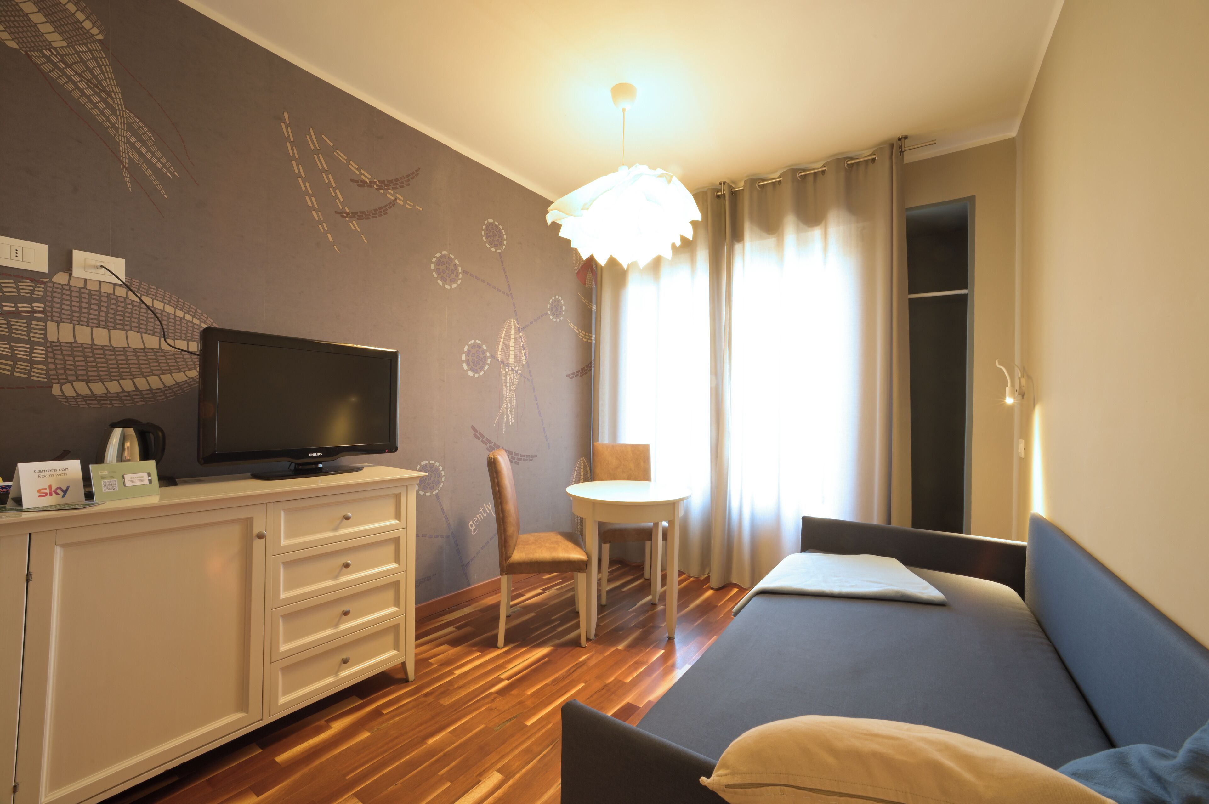 Camera d'albergo moderna con TV a schermo piatto, un piccolo tavolo da pranzo, un divano e un'ampia finestra con tende.