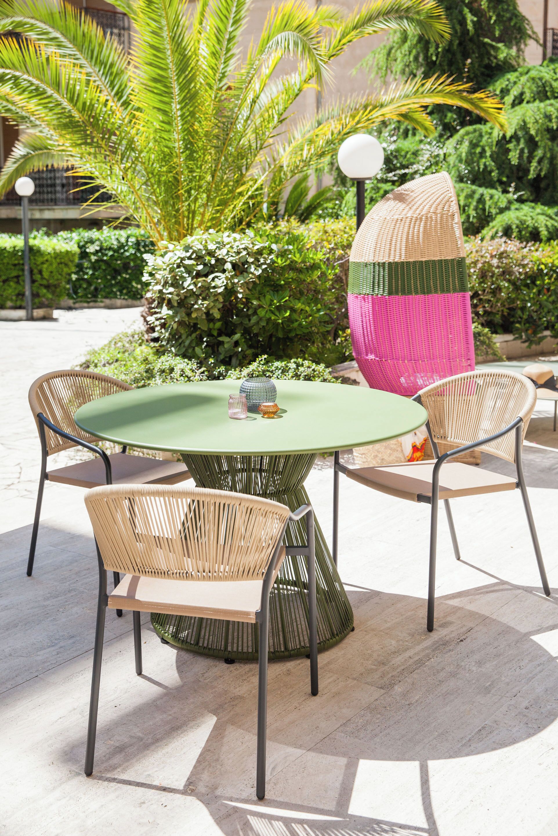 A round table with four wicker chairs, surrounded by lush greenery and a decorative pink and green structure.