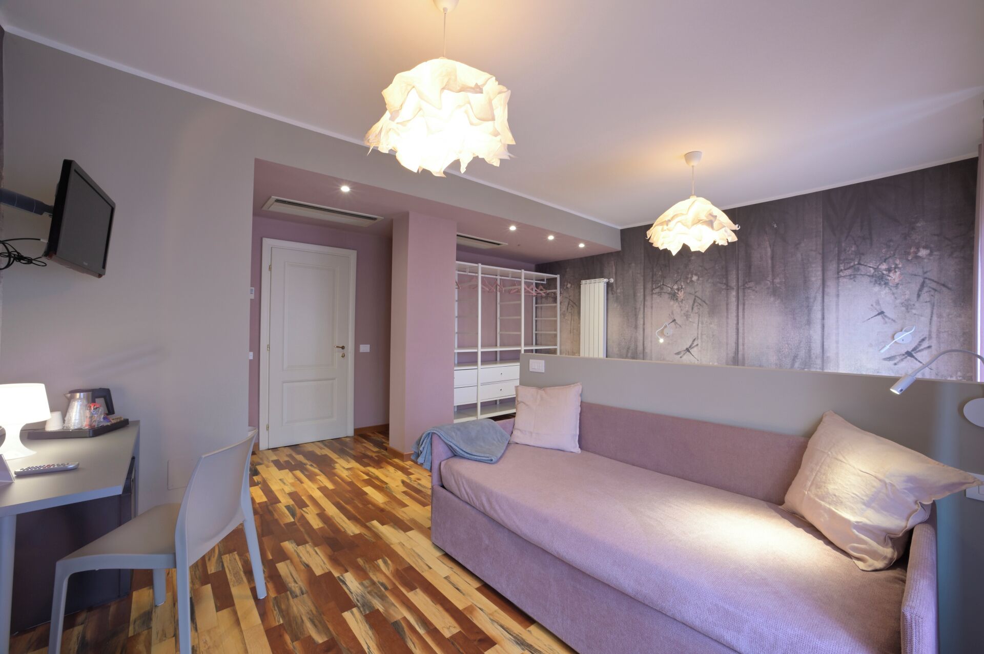 A modern bedroom with a wooden parquet floor, a sofa, a desk with a chair, a wardrobe, and a large mirror.
