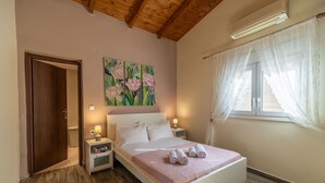 A bedroom with a wooden ceiling, a bed with a pink bedspread, two bedside tables with lamps, a painting of flowers on the wall, and a window with sheer curtains.