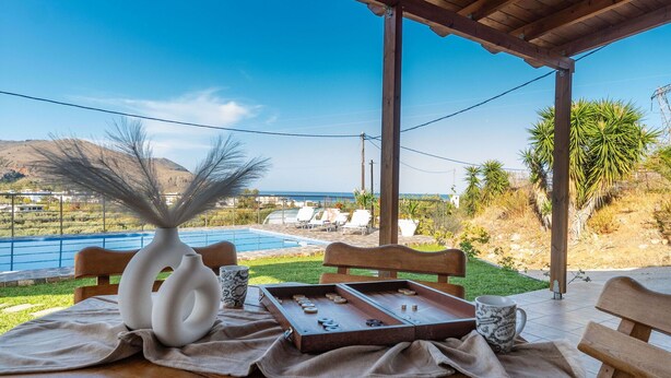 A wooden table with a white vase, a cup, and a board game set on a cloth, overlooking a pool and a coastal view.