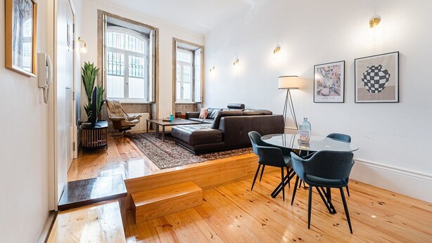 Ein großzügiger Wohnbereich mit einer Couch, einem Couchtisch und einem Esstisch mit Stühlen. Große Fenster sorgen für natürliches Licht und bieten einen Blick nach draußen.