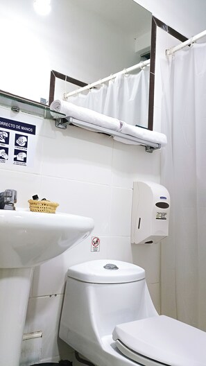 A bathroom with a white sink, toilet, and shower curtain. There is a hand sanitizer dispenser and a basket on the sink. - Di Costa Hoteles (Piura)