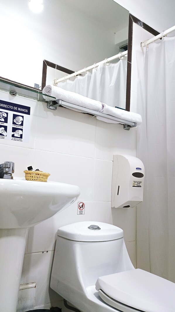 Une salle de bain avec un lavabo, des toilettes et un rideau de douche, tous de couleur blanche. On y trouve un distributeur de gel hydroalcoolique et un panier sur le lavabo.