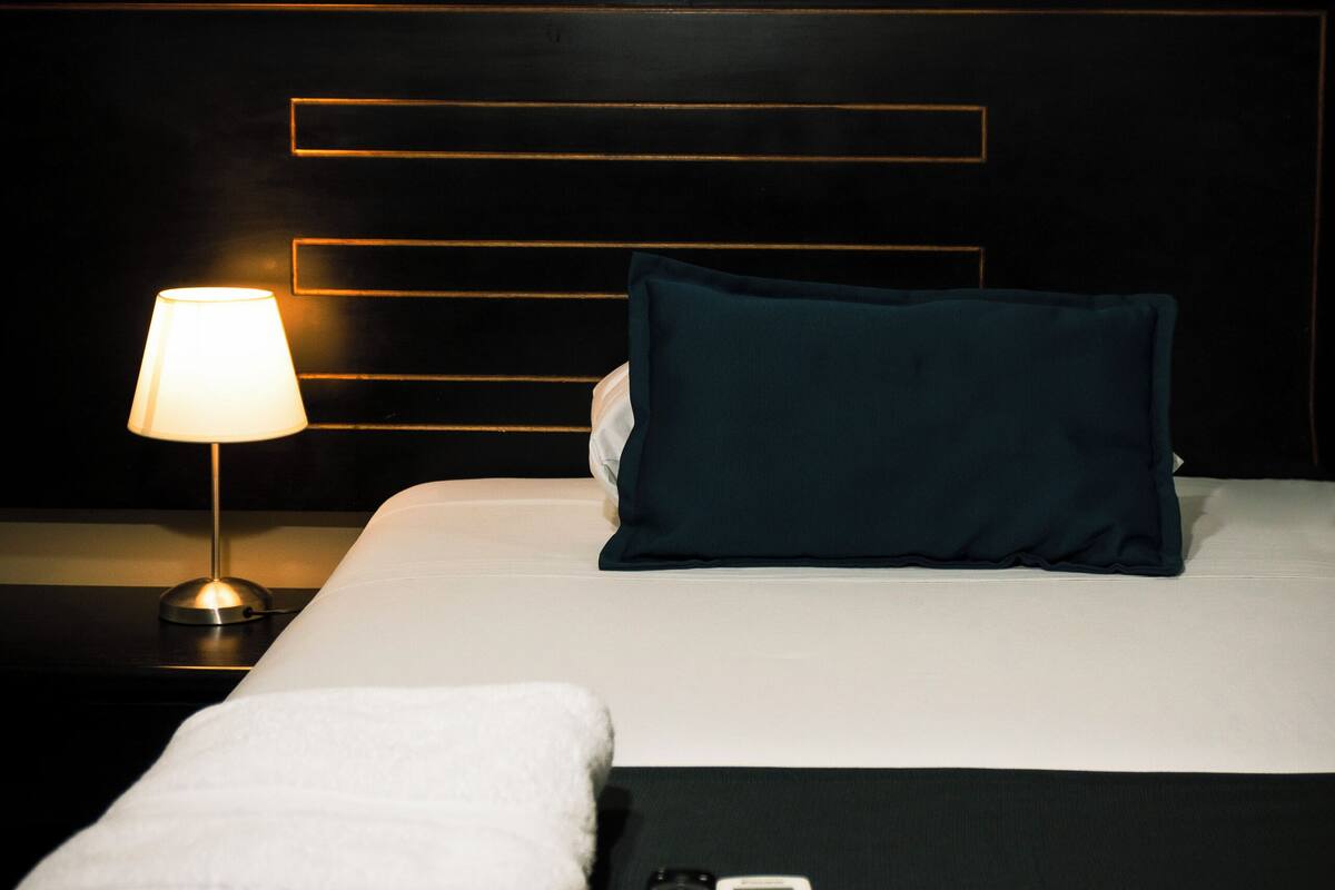 Un lit bien fait, recouvert d’une couverture blanche, avec un oreiller turquoise, une télécommande et une lampe sur une table de chevet.