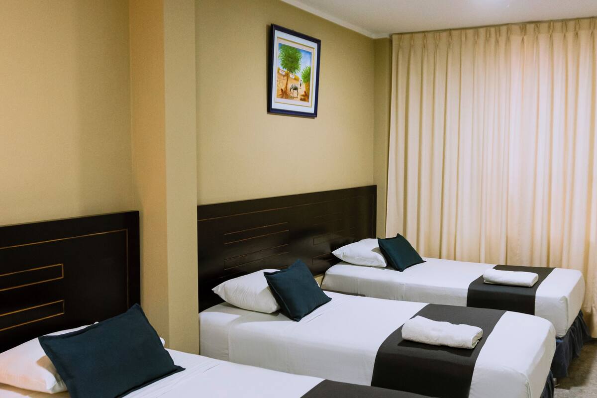 Une chambre d’hôtel avec trois lits simples, chacun doté d’une tête de lit noire et de draps blancs. Un tableau encadré est accroché au mur au-dessus des lits, et il y a une fenêtre avec des rideaux de couleur beige.