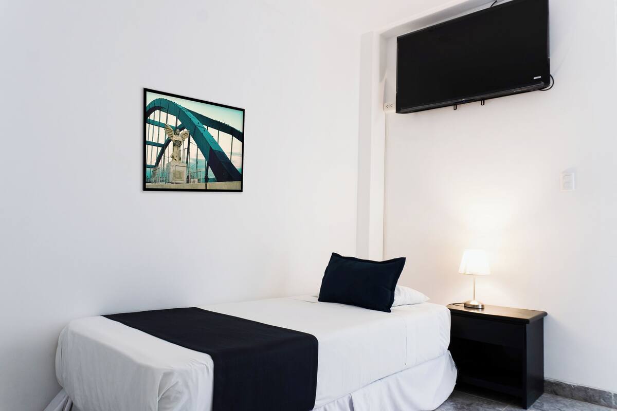 Une chambre d’hôtel avec un lit, une table de chevet avec une lampe, un téléviseur fixé au mur et un tableau représentant un pont.