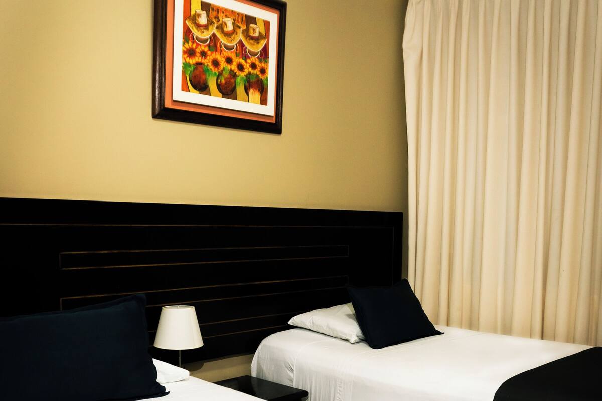 Une chambre d’hôtel avec deux lits, une tête de lit, un tableau encadré au mur et une fenêtre avec des rideaux.