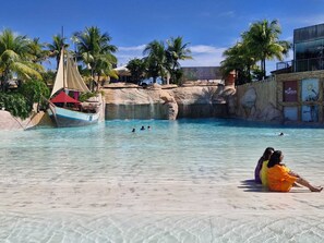 A swimming pool with clear water, a sailboat, palm trees, and a rocky backdrop.