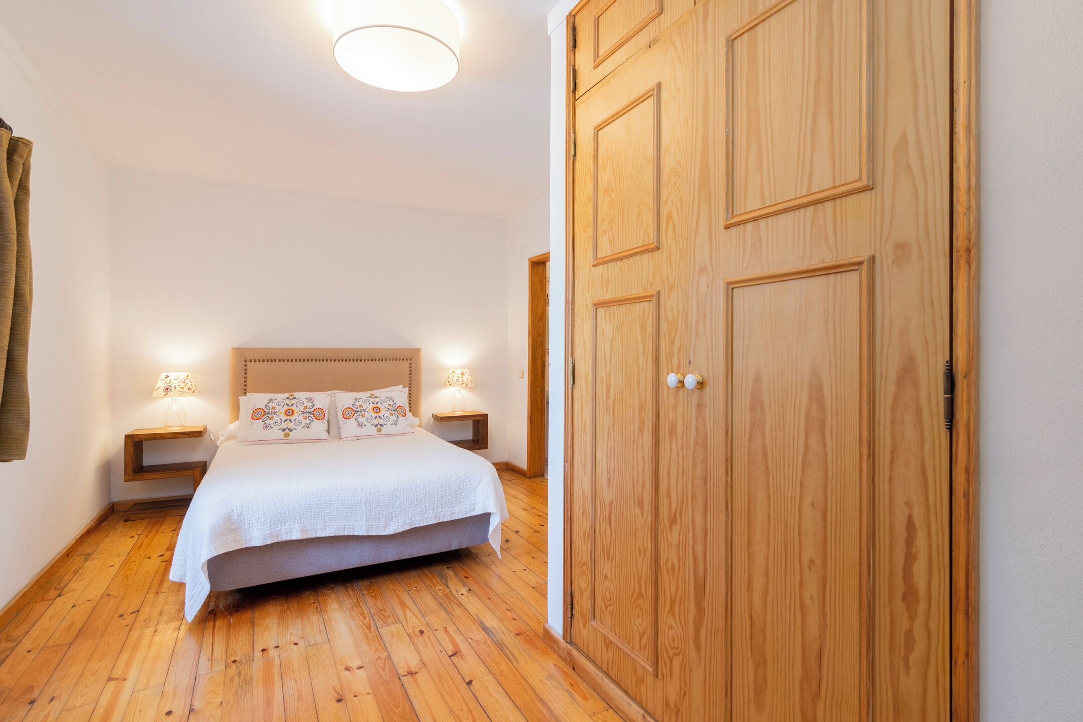 Ein Schlafzimmer mit Holzboden, einem großen Schrank aus Holz, einem Bett mit weißer Bettdecke und gemusterten Kissen sowie Nachttischlampen.