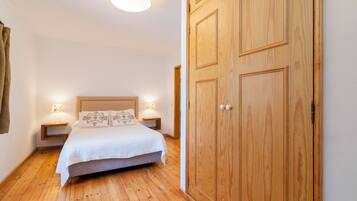 Quarto com piso de madeira, um guarda-roupa grande de madeira, cama com colcha branca e almofadas estampadas, e abajures laterais.