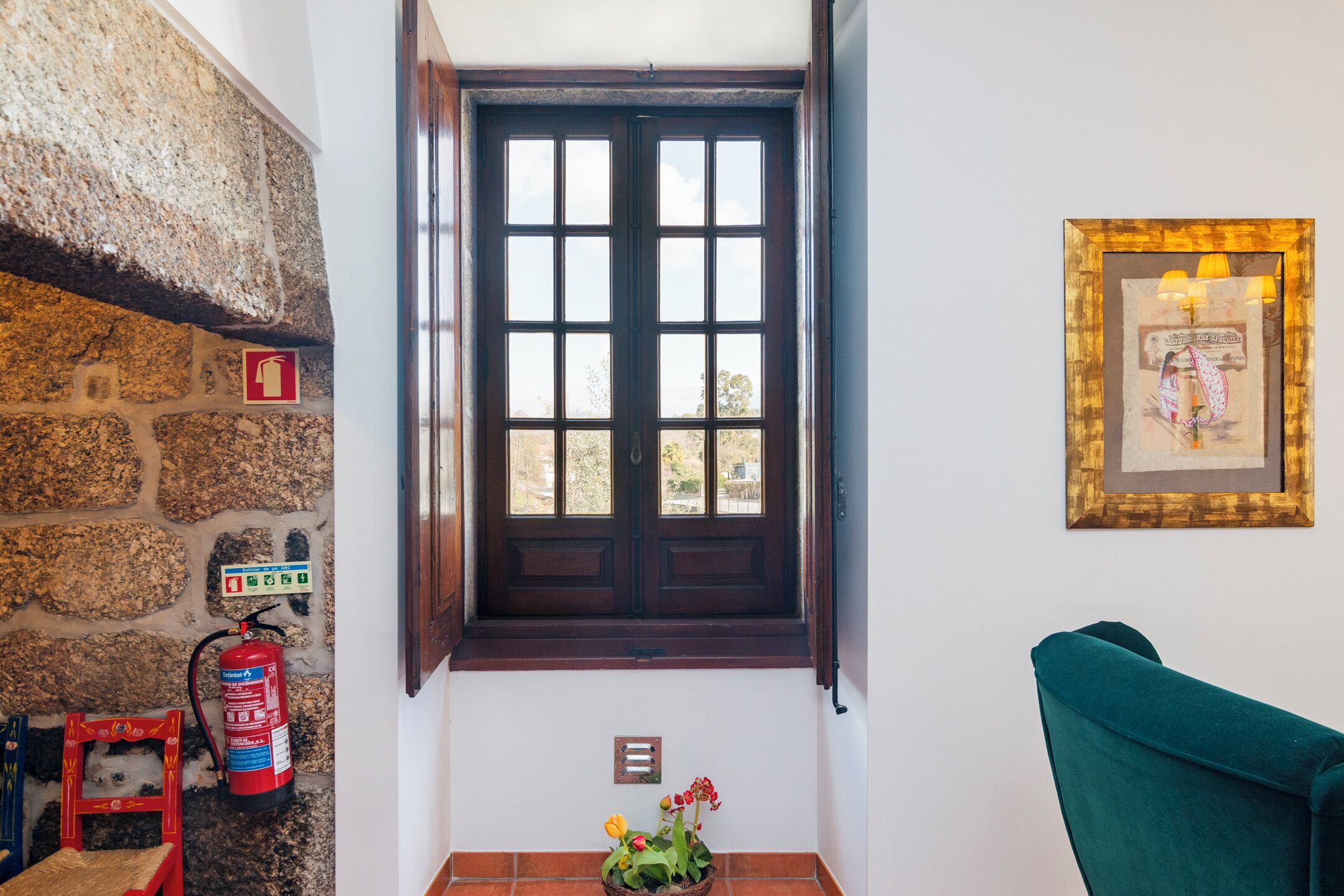 A room with a stone wall, a wooden door, a fire extinguisher, a small stool, a chair, and a framed picture on the wall.