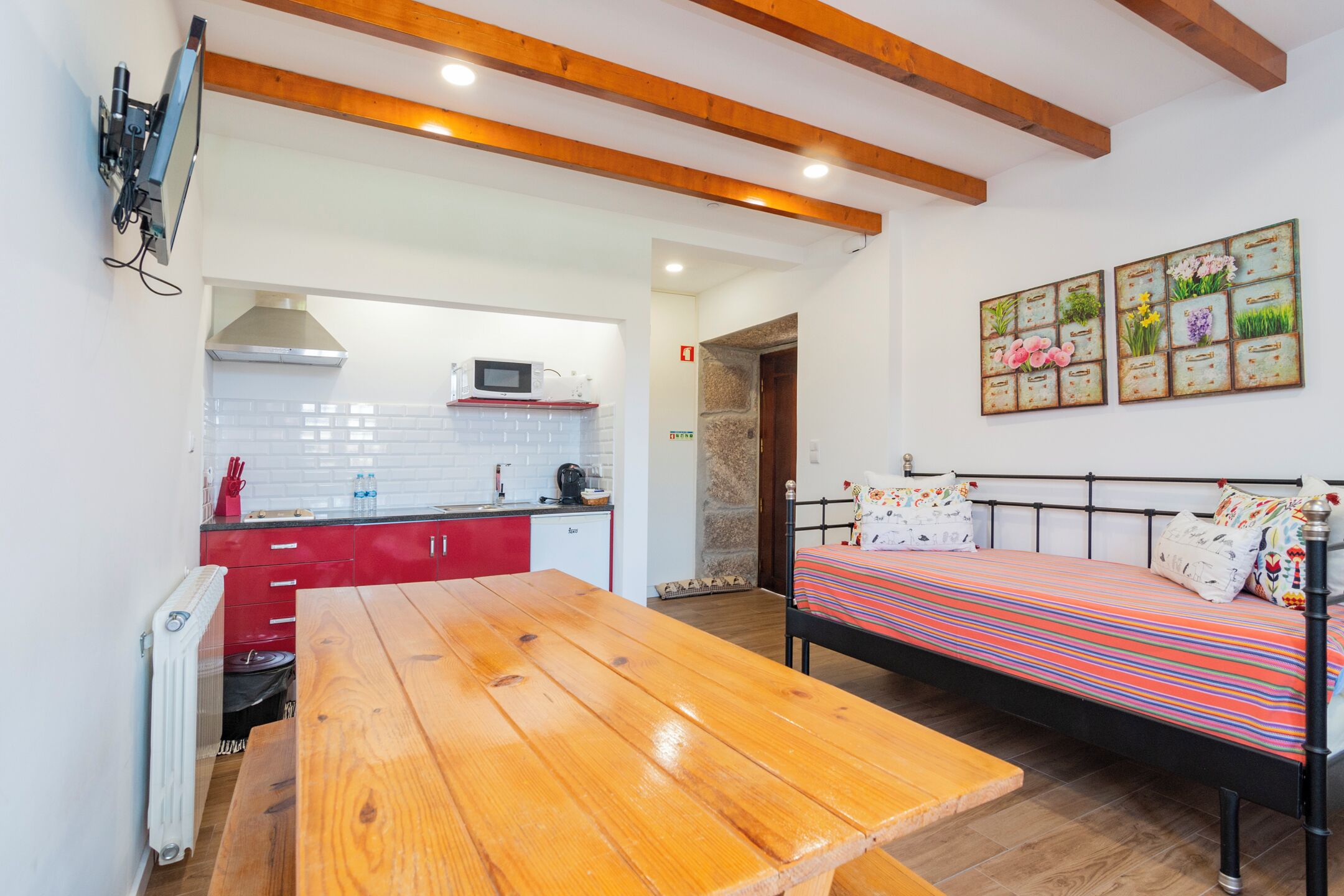 A compact room with a wooden table, a bed with a colorful striped bedspread, a kitchenette with a microwave, and a red cabinet.