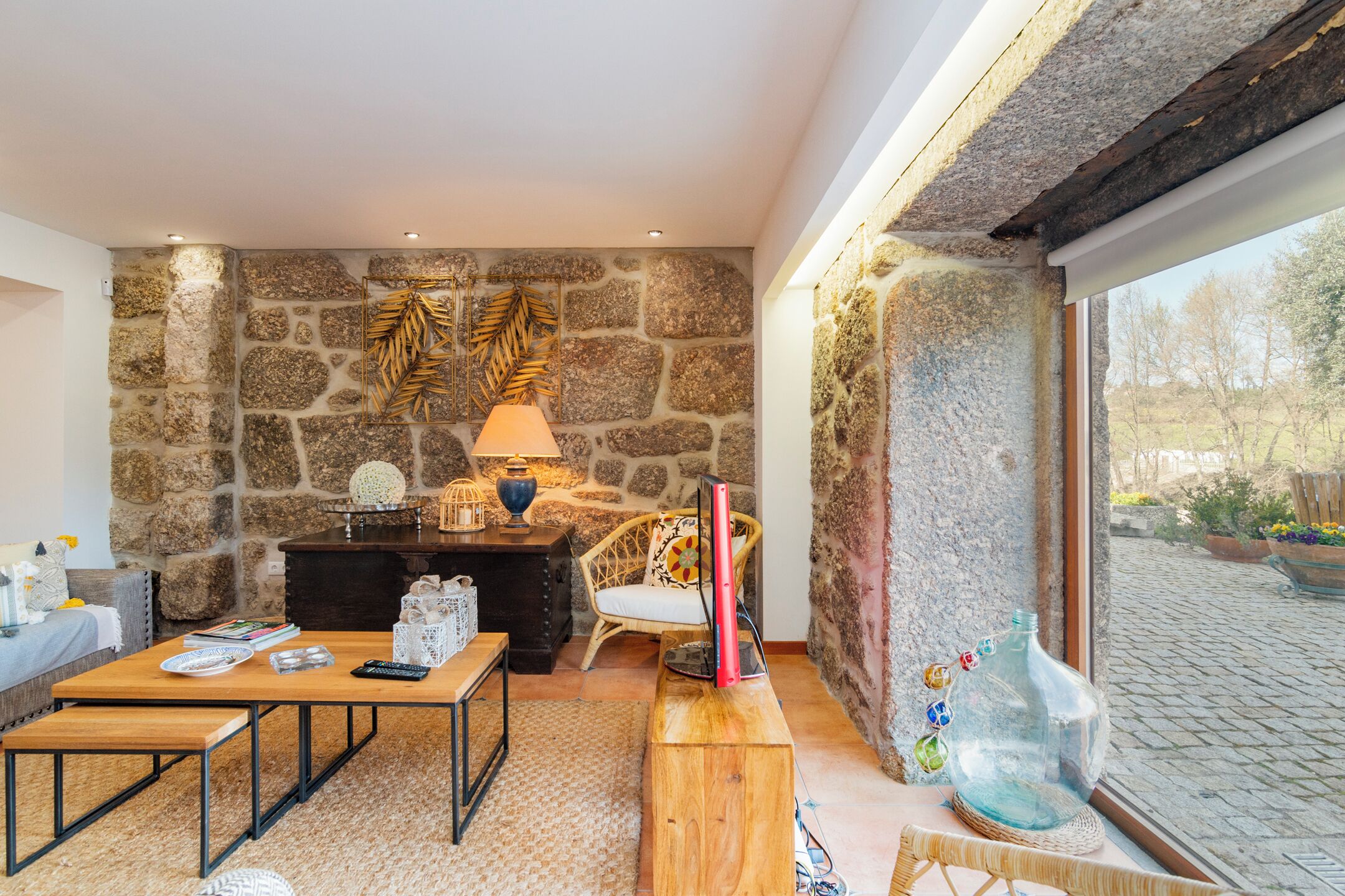 A cozy living room with a stone wall, a wooden table, a comfortable chair, and a large window offering a view of the outdoors.