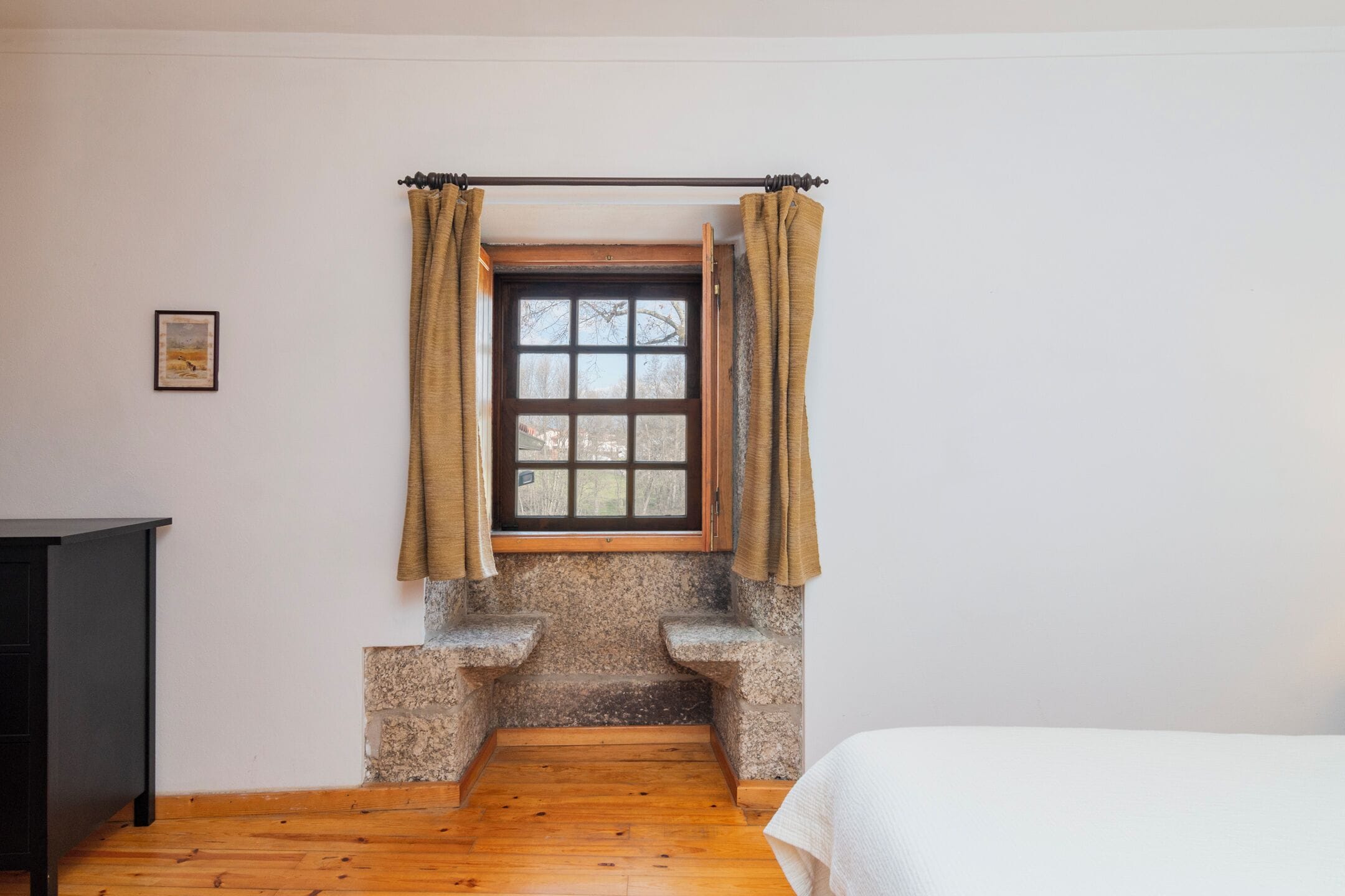 Una habitación con un banco de piedra, una ventana con cortinas, un cuadro en la pared y una cama.