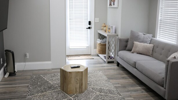 Una sala de estar con un sofá gris, una mesa de centro de madera, una puerta blanca con persianas y un estante fijado a la pared con objetos decorativos.