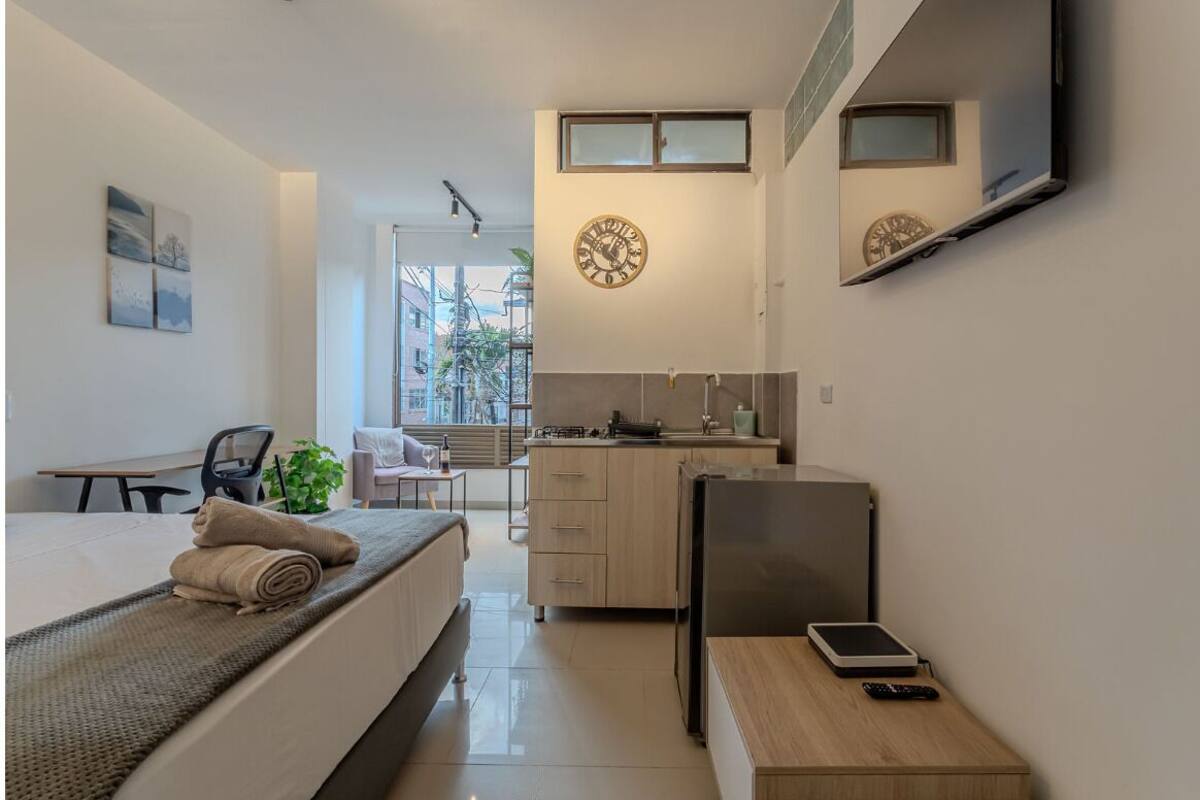 A compact hotel room with a bed, a desk, a small kitchenette, and a TV mounted on the wall.