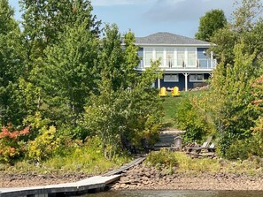 A house with a dock extending into a body of water, surrounded by trees and greenery. - Lakefront Home on Washademoak Lake with Dock and 255 ft of Water Frontage.     (Coles Island)