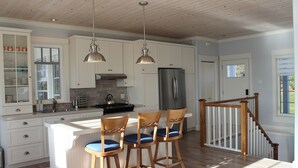 A kitchen with white cabinets, a stainless steel refrigerator, and wooden bar stools with blue cushions. - Lakefront Home on Washademoak Lake with Dock and 255 ft of Water Frontage.     (Coles Island)