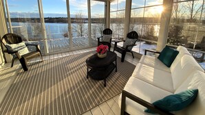 A sunlit room with a white sofa, two black chairs, a small table, and a large window overlooking a snowy landscape. - Lakefront Home on Washademoak Lake with Dock and 255 ft of Water Frontage.     (Coles Island)