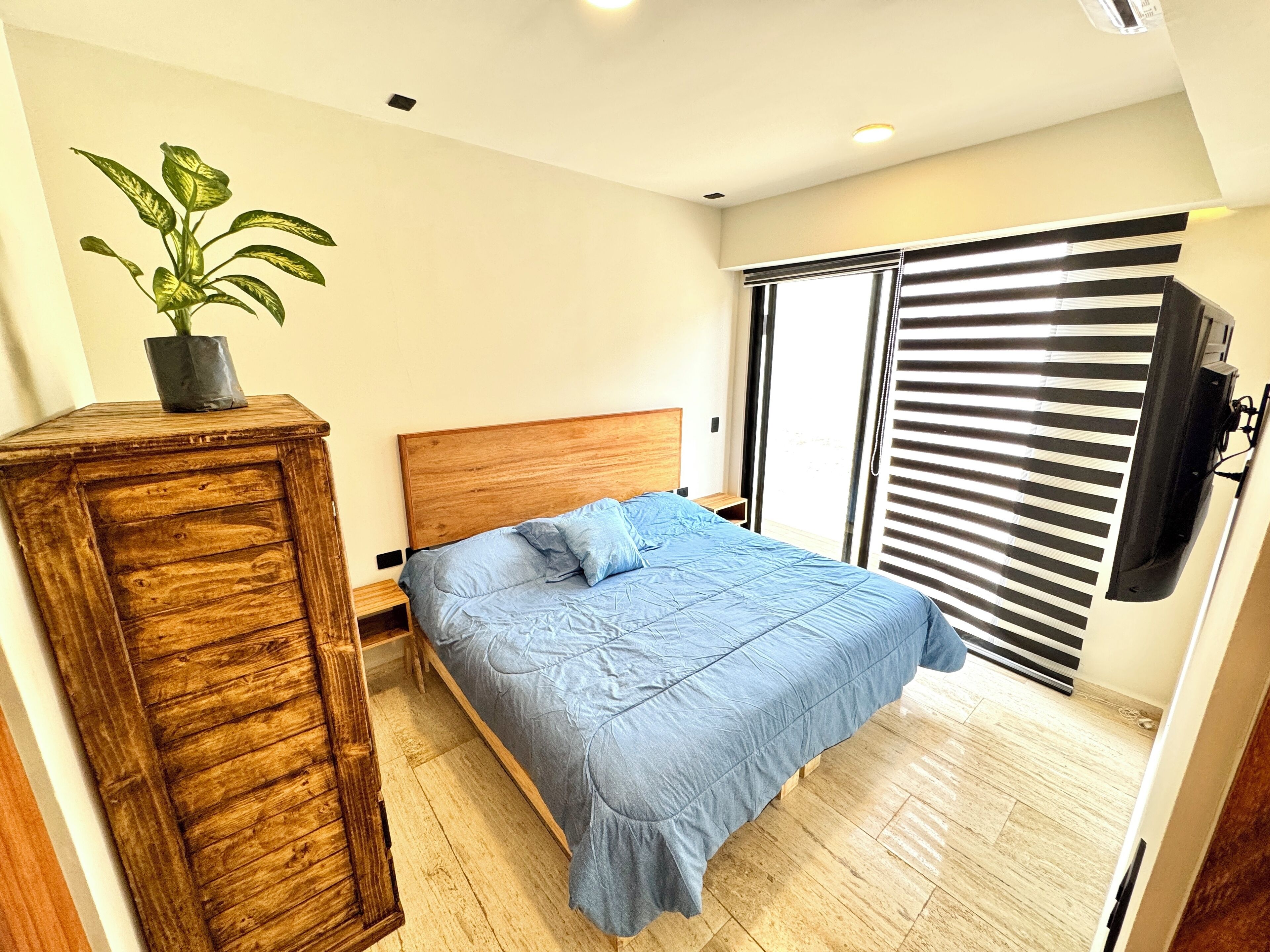 A bedroom with a bed, a wooden dresser, a potted plant, a TV, and a sliding door with blinds.