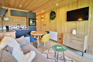Uma sala de estar aconchegante com um sofá, uma mesa de jantar com cadeiras, uma cozinha com armários e eletrodomésticos, e uma TV fixada na parede.
