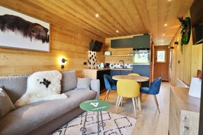 Interior acolhedor de uma cabana com sofá, mesa de jantar com cadeiras, área de cozinha e teto de madeira.
