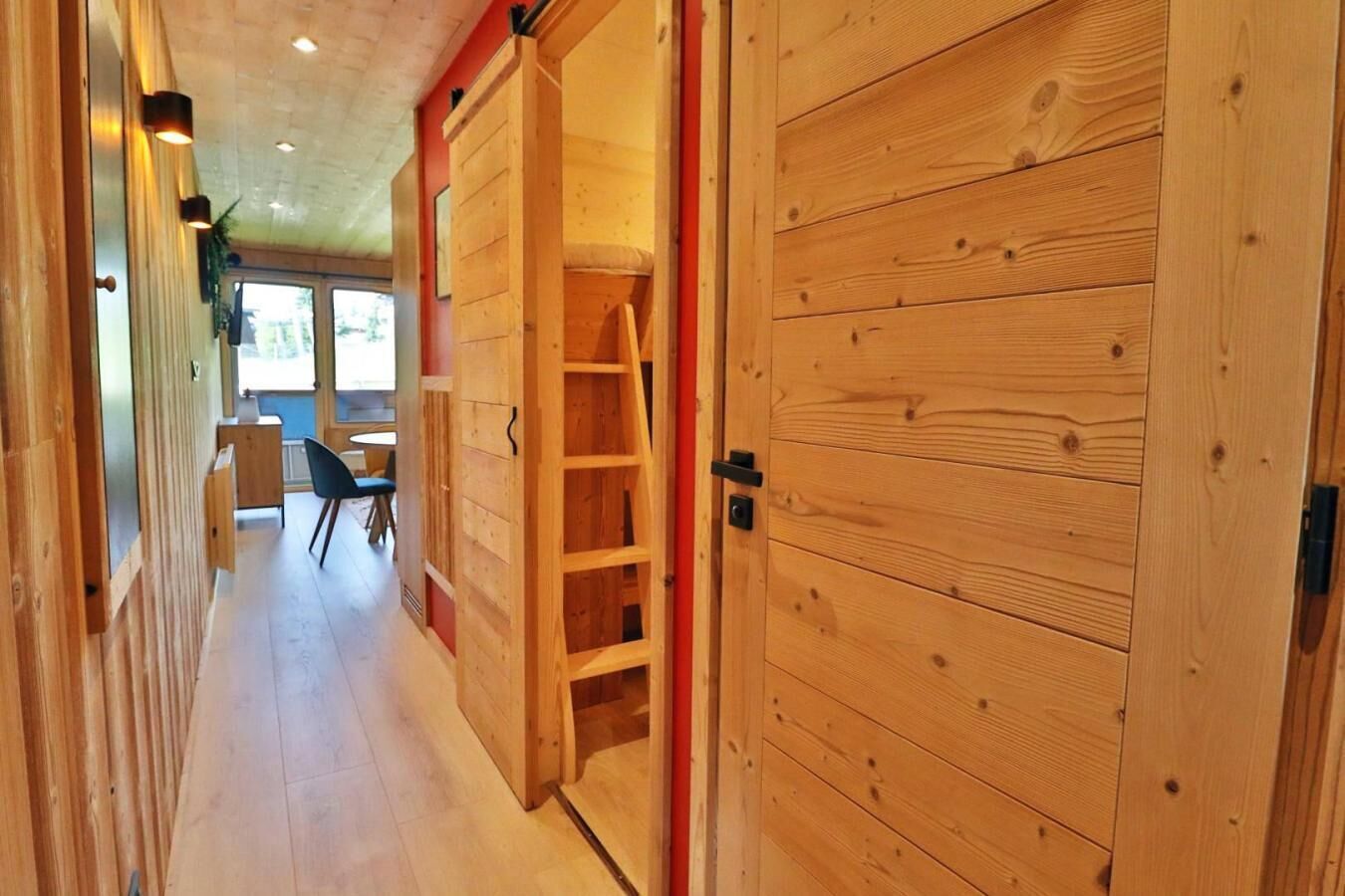 A wooden interior hallway with a sliding door, a ladder, and a view into a dining area with chairs and a table.