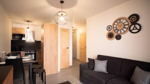 Een moderne woonkamer met een donkergrijze bank, een klein bijzettafeltje en een eethoek met een tafel en stoelen. In de achtergrond is een keuken met een magnetron en kookplaat zichtbaar.