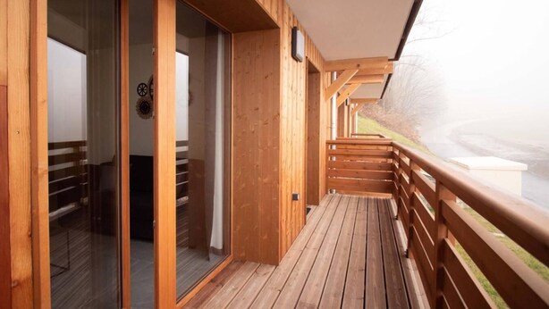 Een houten terras met een glazen deur die leidt naar een kamer, een houten leuning en uitzicht op een wateroppervlak.