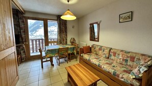 A cozy living room with a wooden sofa, a wooden coffee table, and a dining table with chairs. There is a window with a view of mountains.