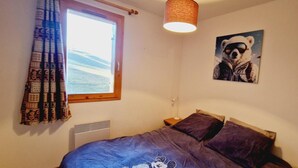 Ett sovrum med en säng, ett fönster med gardiner, en vägglampa, ett element och en affisch med en björn på.