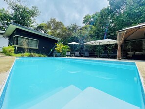 A rectangular swimming pool with a tiled edge, surrounded by outdoor seating with umbrellas, a building with a dark facade, and lush greenery in the background. - Escápate al paraíso Domos con jacuzzi y terraza privada dentro de la naturaleza (Cóbano)