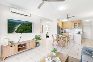 A modern living room with a flat-screen TV, wooden entertainment unit, dining area with chairs, and a kitchen with cabinets and appliances.