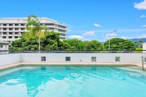 A swimming pool with a clear blue water surface, surrounded by a white building with multiple floors and balconies, and lush green trees in the background.