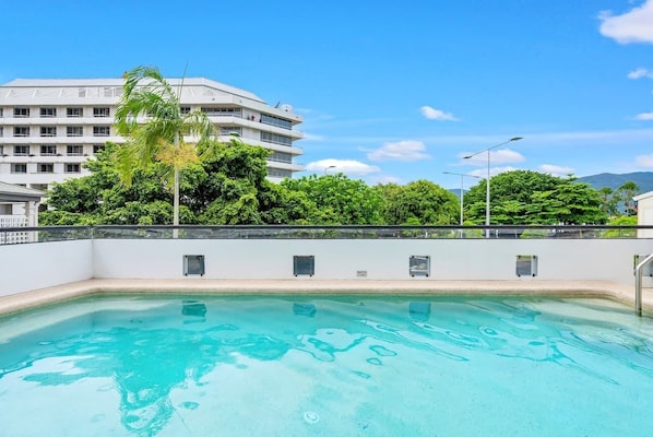 Una piscina con superficie d'acqua azzurra e cristallina, circondata da un edificio bianco con diversi piani e balconi, e alberi verdi rigogliosi sullo sfondo.